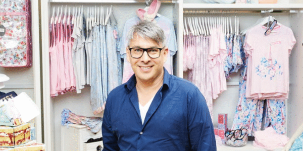 Peter Alexander, wearing a navy shirt and glasses, smiles in front of racks of pastel sleepwear inside a brightly lit Peter Alexander store.